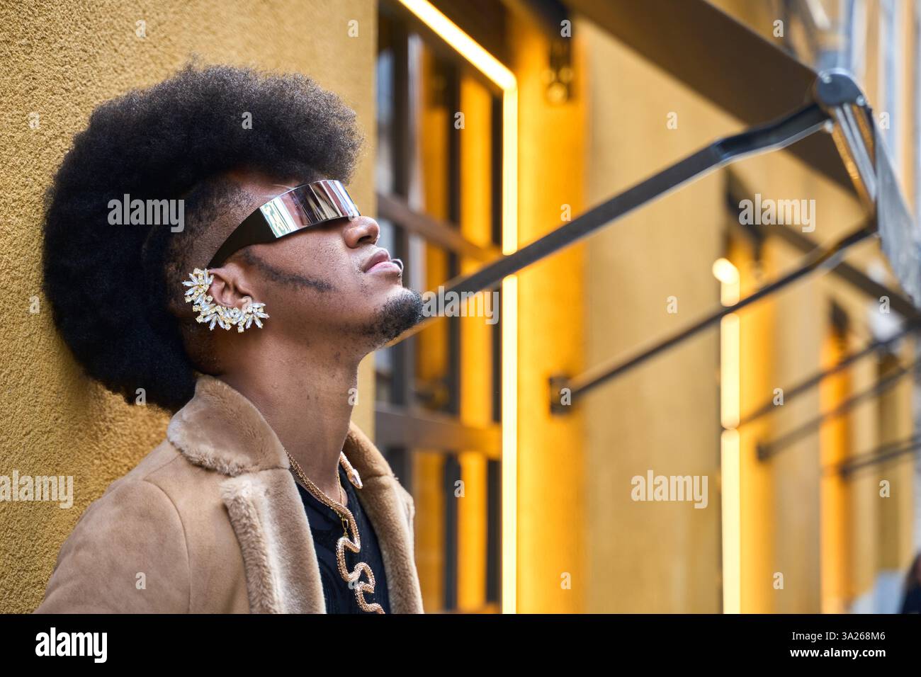 Elegante persona androgina con acconciatura afro e occhiali da sole futuristici in posa vicino alle pareti gialle in un ambiente urbano Foto Stock