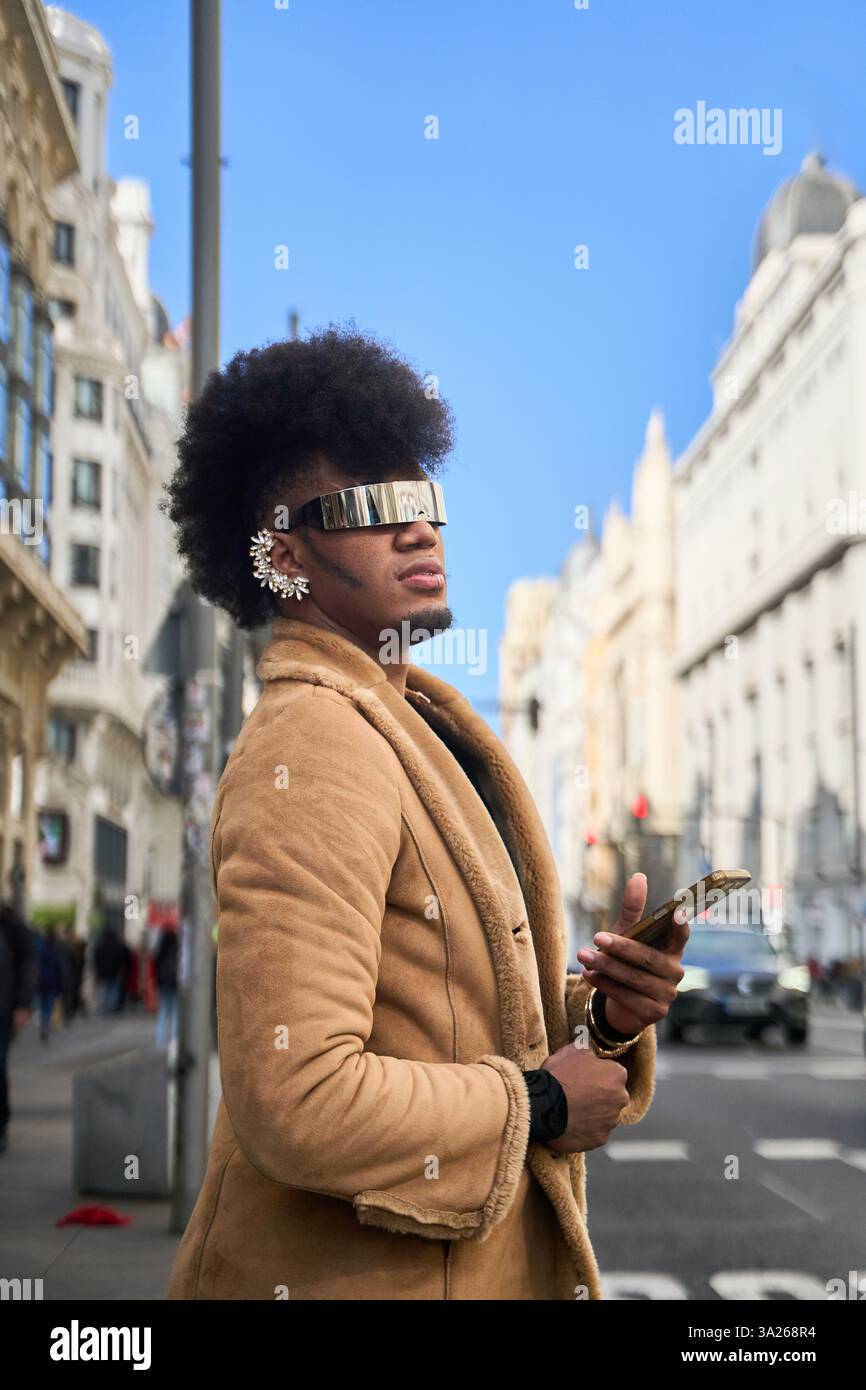Persona androgina che indossa occhiali da sole e orecchini futuristici e tiene in mano uno smartphone mentre cammina in una strada della città Foto Stock
