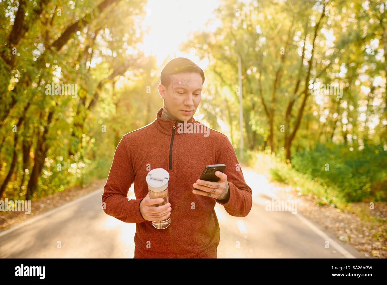 Un giovane con i tatuaggi percorre un sentiero panoramico, beve in mano, controlla il suo telefono. Foto Stock