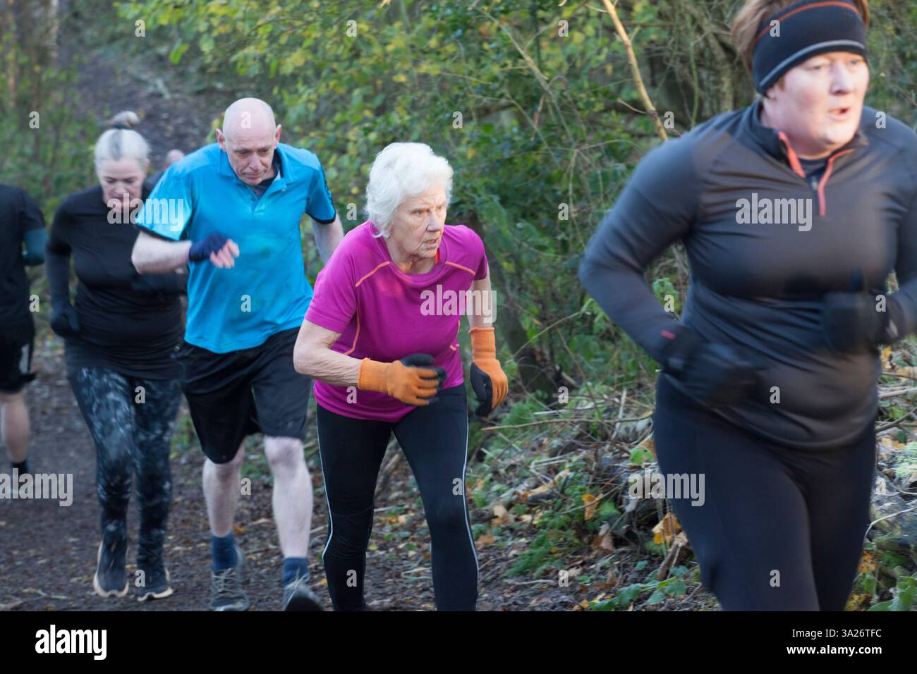 Un gruppo di persone che fanno jogging in salita su un sentiero nella foresta, vestite con abbigliamento atletico adatto al clima fresco. Sale Water Park, Manchester, Regno Unito Foto Stock