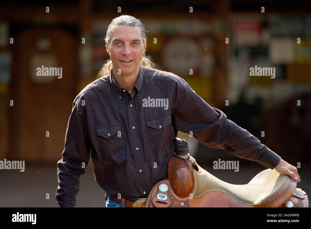 L'uomo con i capelli lunghi in una camicia scura si appoggia su una sella e sorride all'aperto. Brandeburgo, Germania Foto Stock