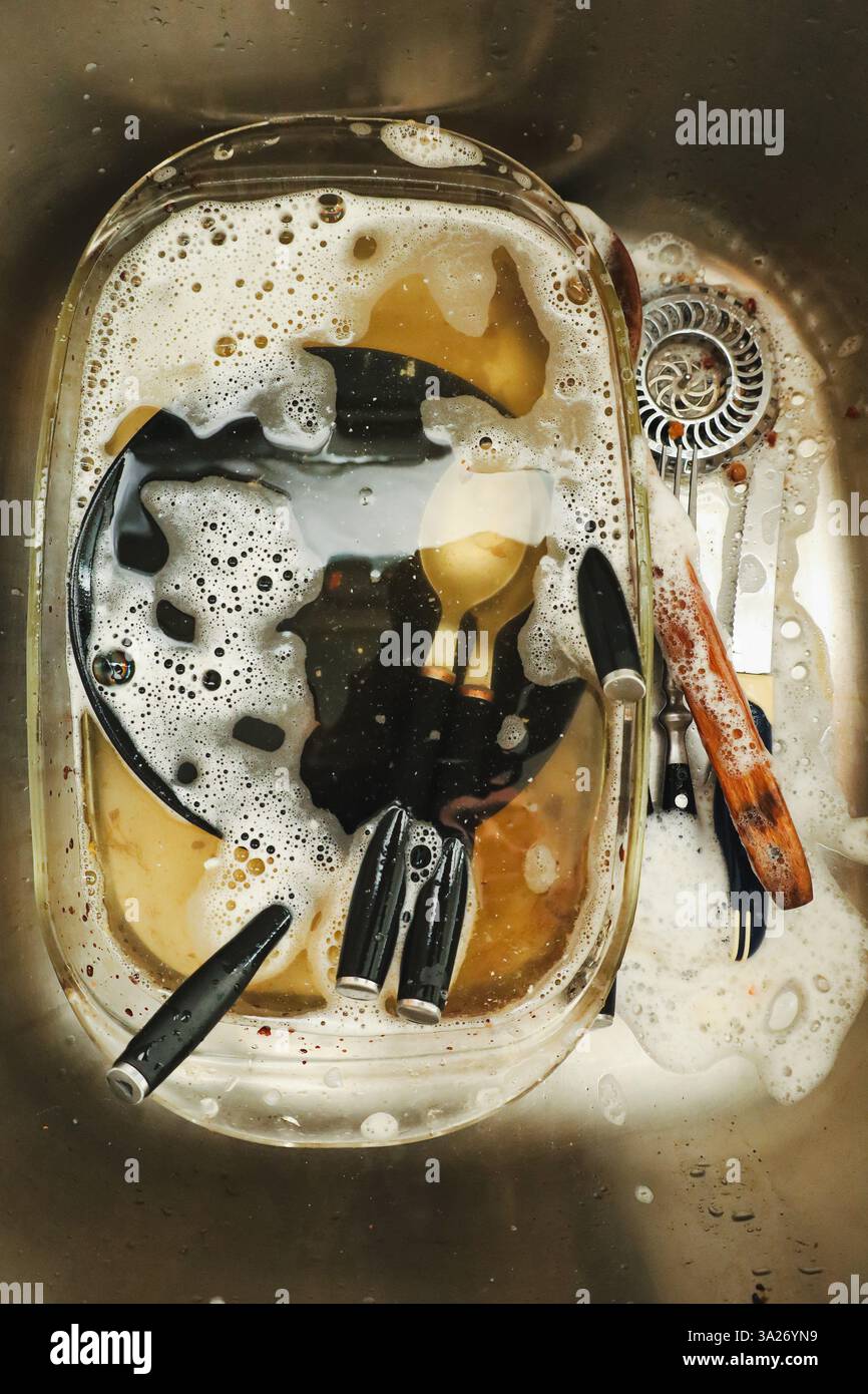 Una vista ravvicinata di un lavandino in acciaio inossidabile pieno di piatti sporchi immersi in acqua e sapone, un compito universale di lavare i piatti Foto Stock