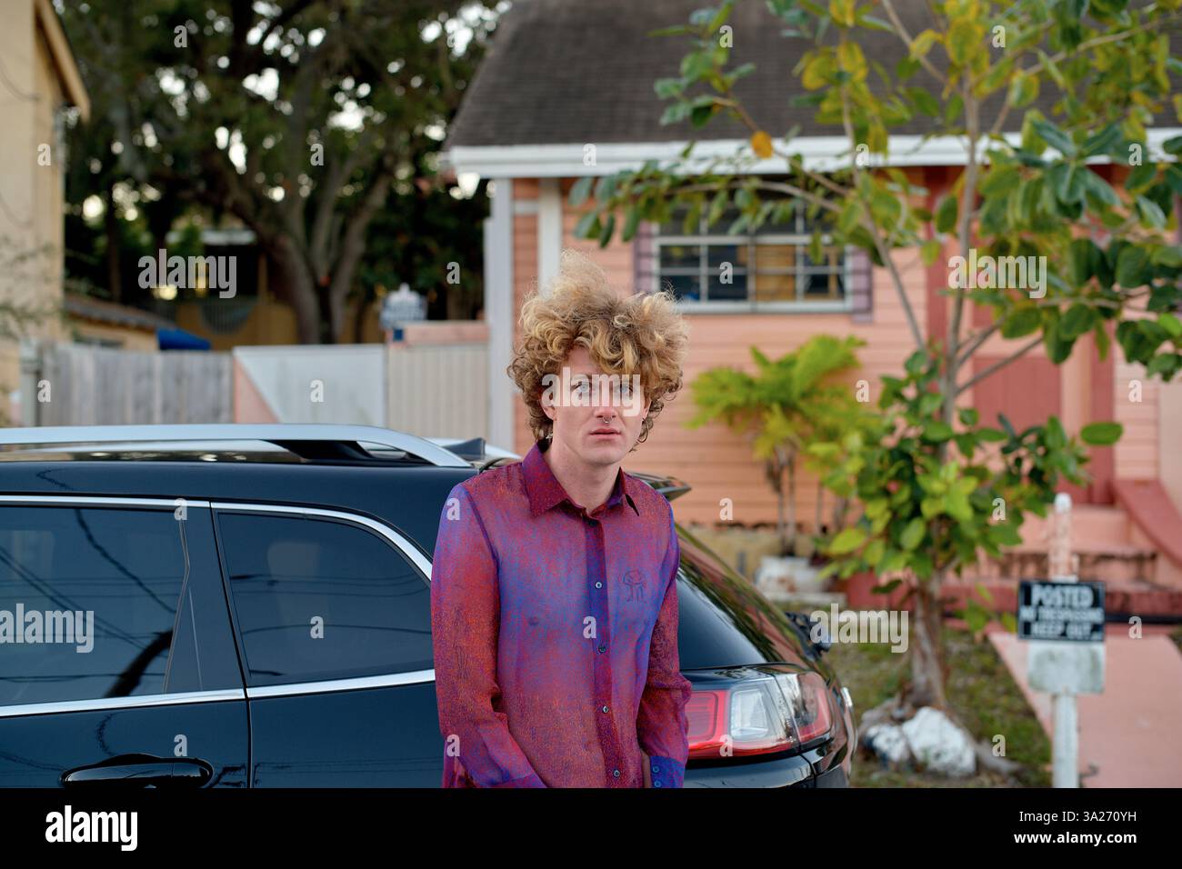 Un giovane con i capelli ricci si trova accanto a un'auto nera di fronte a una casa color pesca. Miami, Florida, Stati Uniti Foto Stock