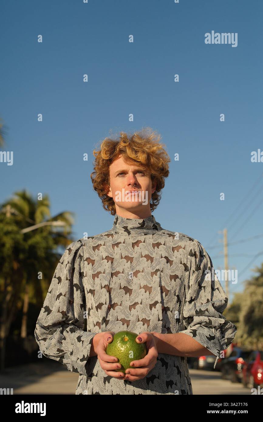 Giovane con i capelli ricci in un vestito che tiene un frutto verde sotto un cielo azzurro. Miami, Florida, Stati Uniti Foto Stock