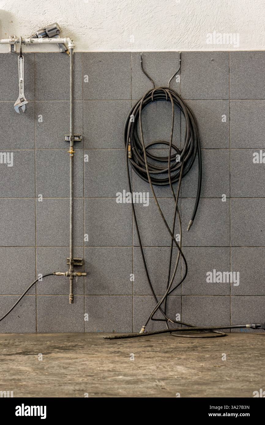 Un tubo flessibile nero dell'aria pressurizzata appeso a una parete, scollegato dal sistema dell'aria, e una chiave regolabile laterale, in un'officina automobilistica. Foto Stock