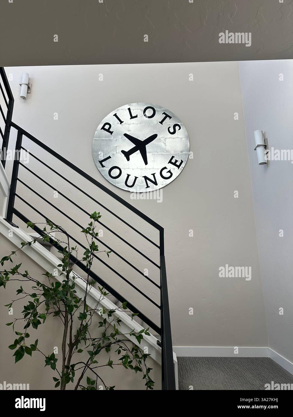 Un elegante cartello Pilots Lounge con una grafica aeroplano è appeso sopra una moderna scala, accentuata da una pianta in vaso e da una soffice illuminazione a parete. - Immagine stock catturata con smartphone