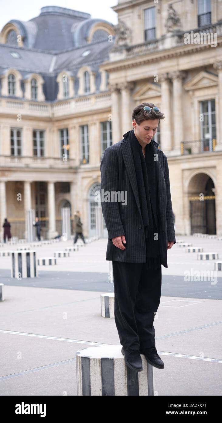Un uomo alla moda in un abito scuro si erge su una colonna a righe al Palais-Royal di Parigi, che mostra eleganza, equilibrio e stile moderno in un'architettura storica. Foto Stock