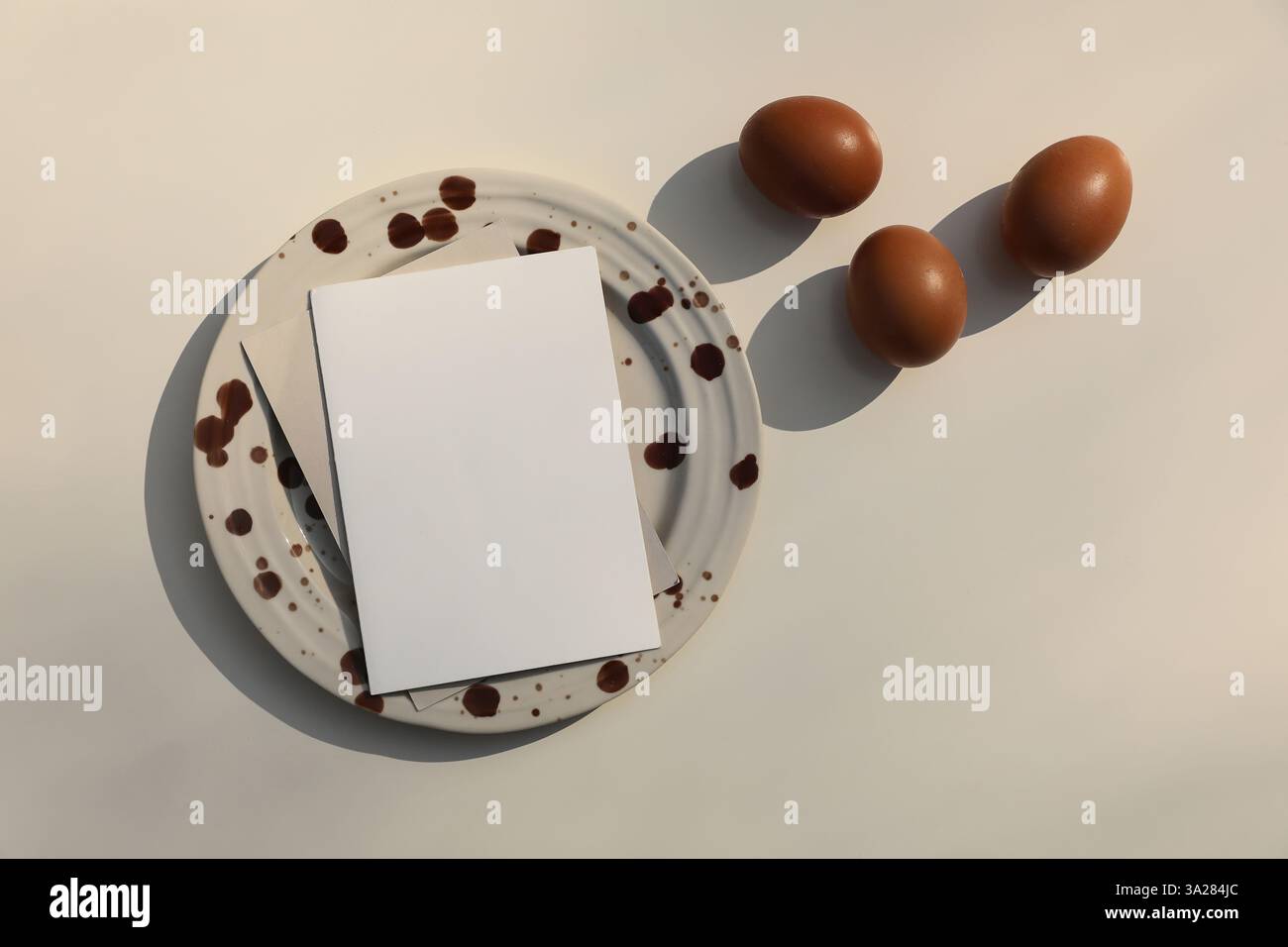La natura morta di Pasqua. Piatto in ceramica macchiata marrone con biglietto d'auguri vuoto, invito. Uova di gallina sullo sfondo del tavolo beige alla luce del sole, ombre lunghe Foto Stock