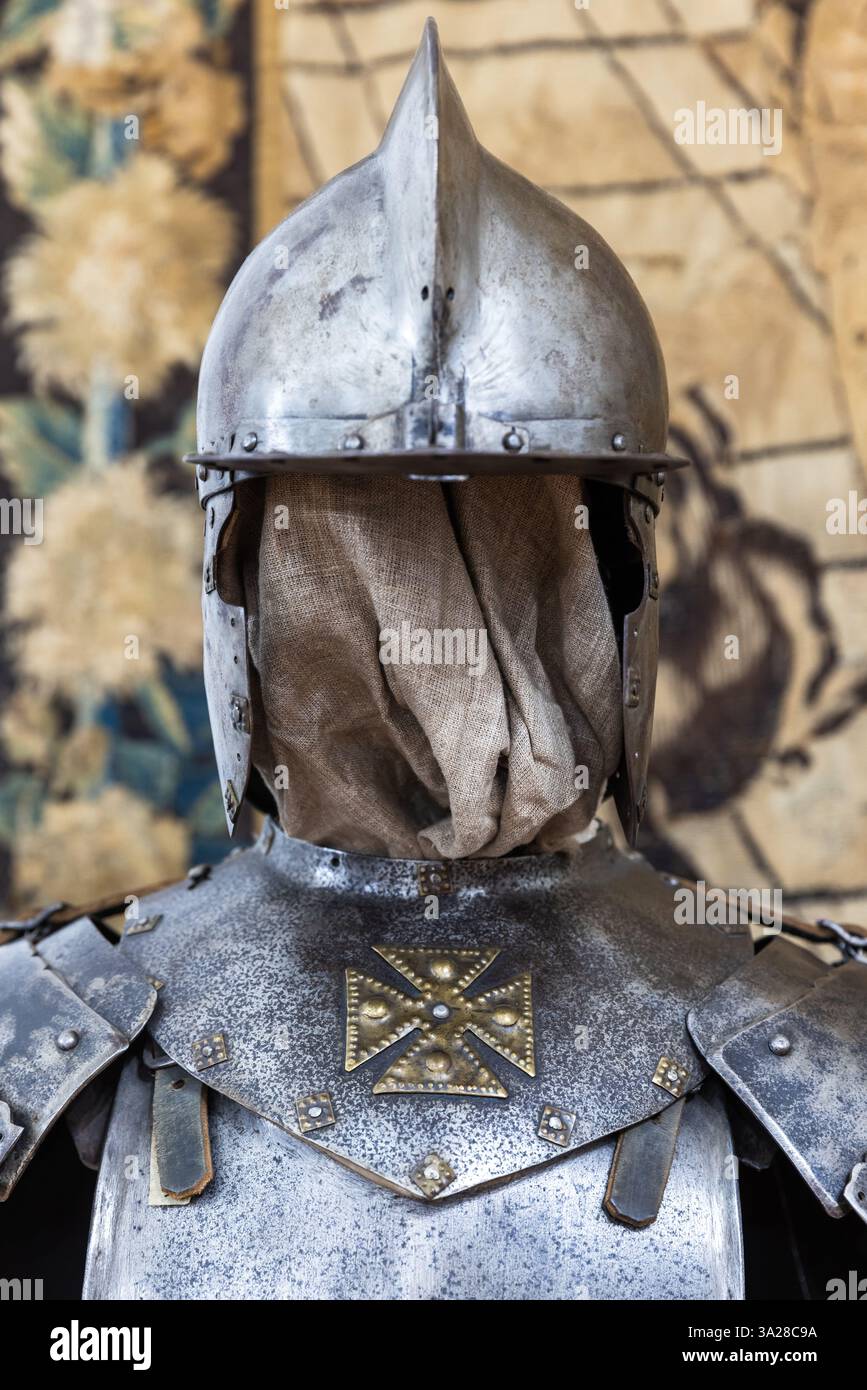 Armatura da cavaliere con casco in acciaio e croce decorativa, catturata su uno sfondo arazzo storico, che mostra l'artigianato medievale e l'herita Foto Stock