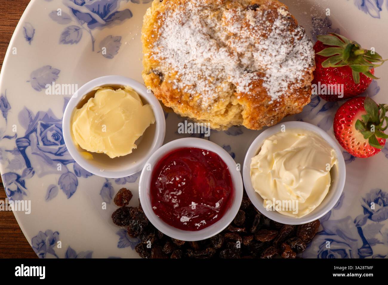 Un piatto di cibo con scone con tè alla panna e condimenti. Il dessert è uno scone con sopra zucchero a velo. I condimenti includono marmellata di fragole e bu Foto Stock