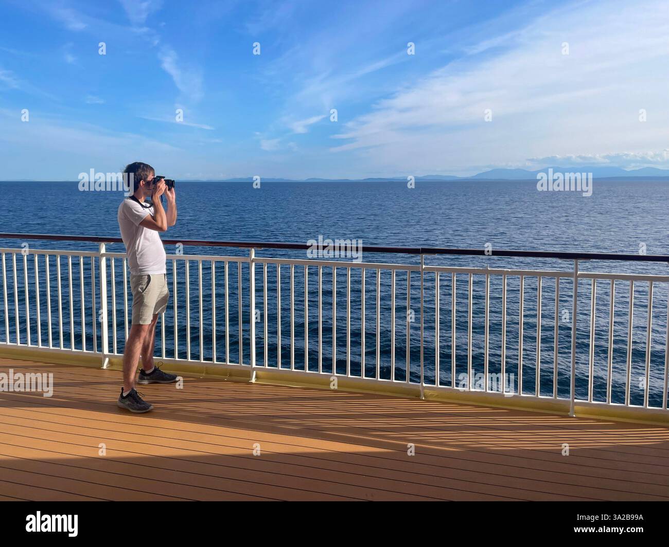 Un uomo che usa un binocolo sul ponte di una nave da crociera, che guarda l'oceano e le montagne lontane. Foto Stock