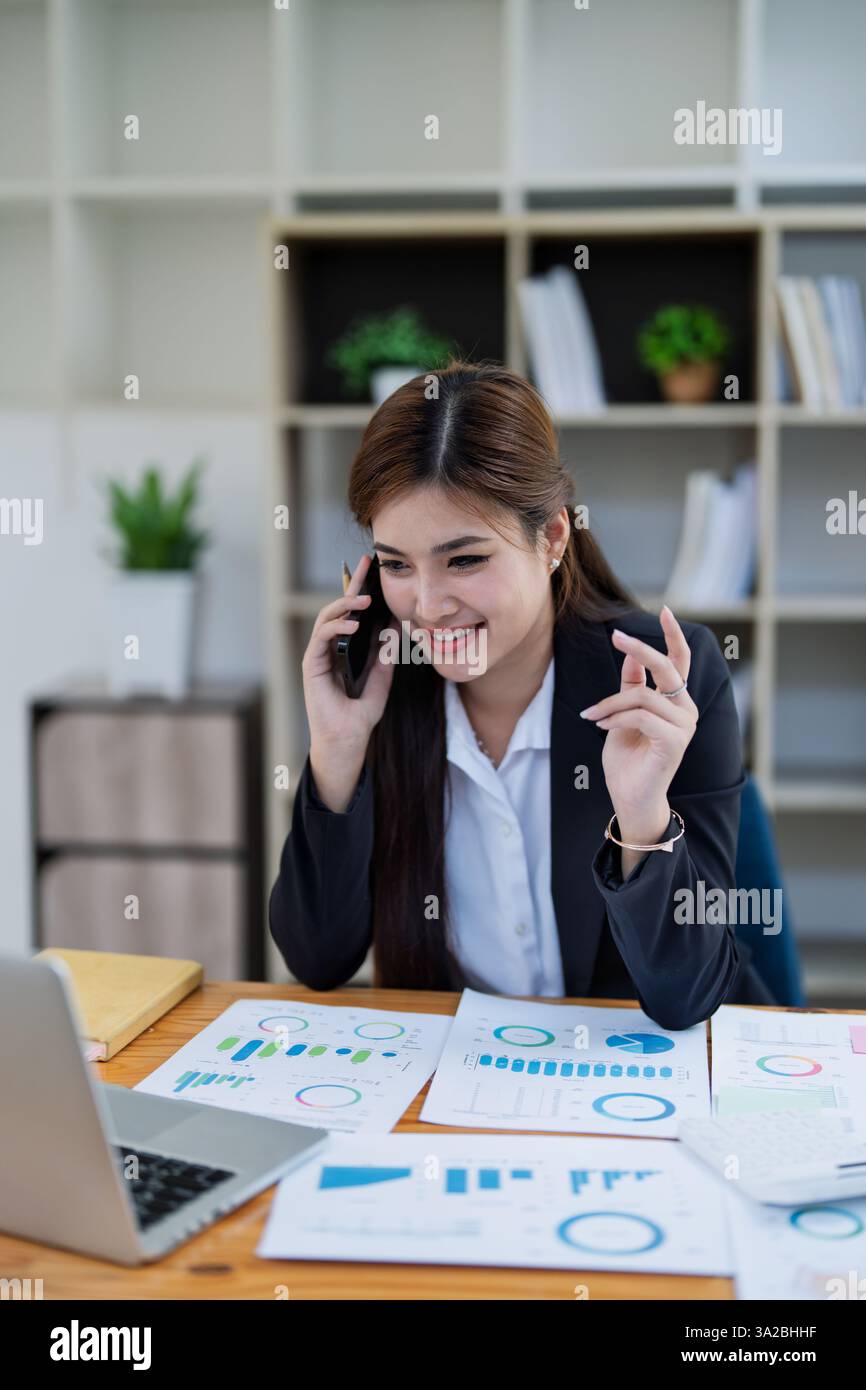 Donna d'affari professionista impegnata in una telefonata tramite smartphone tramite calcolatrice per calcolare la finanza utilizzando grafici di stock marketing, notebook e laptop in of Foto Stock