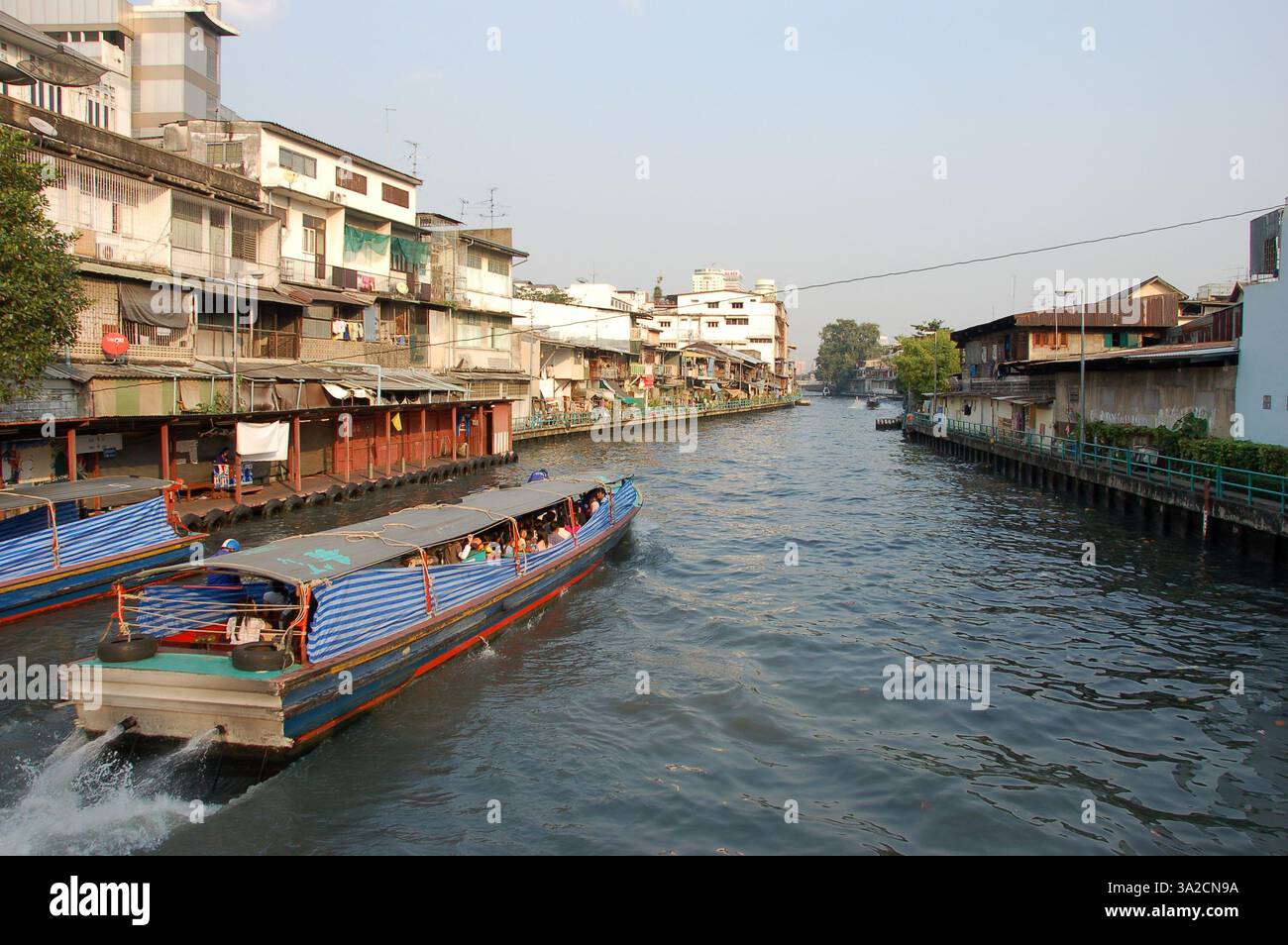 I taxi d'acqua navigano su un canale di Bangkok, con tradizionali palafitte e vita locale visibili lungo il corso d'acqua sotto un cielo limpido Foto Stock