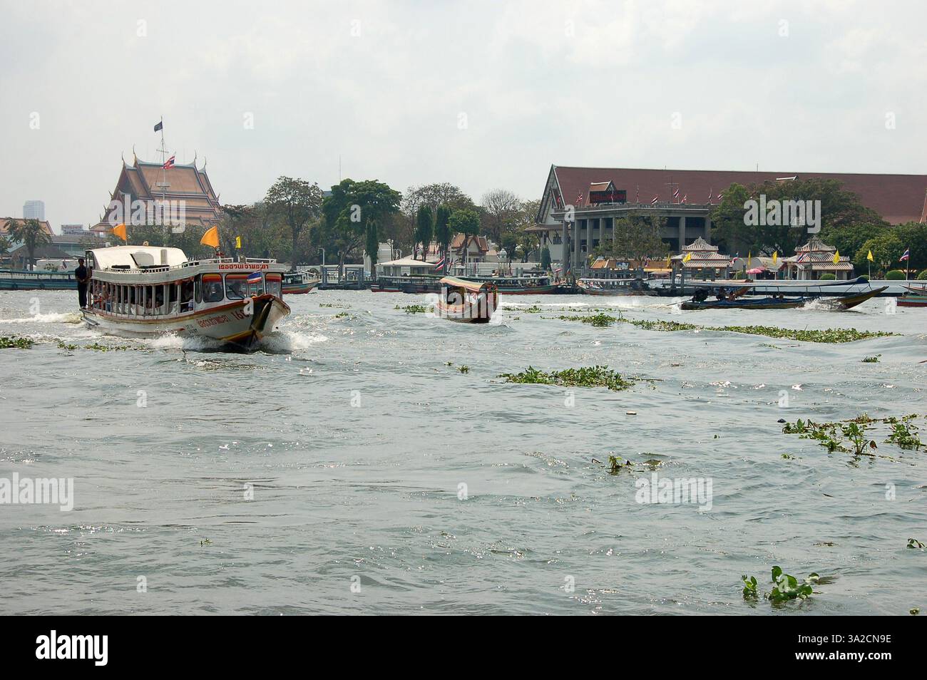 I taxi d'acqua navigano su un canale di Bangkok, con tradizionali palafitte e vita locale visibili lungo il corso d'acqua sotto un cielo limpido Foto Stock
