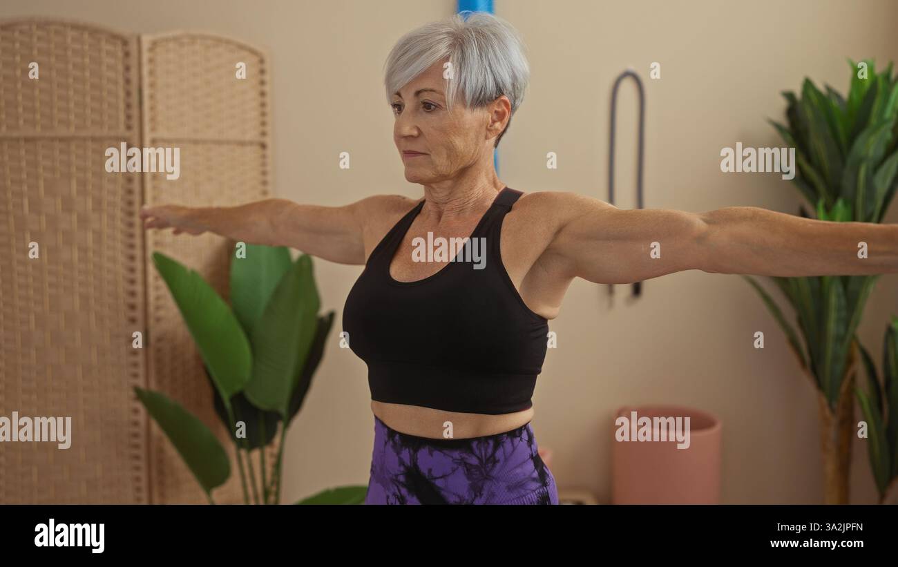 Bella donna anziana con i capelli grigi corti che si allena in palestra, indossa un top nero e leggings viola, allungando le braccia. Foto Stock