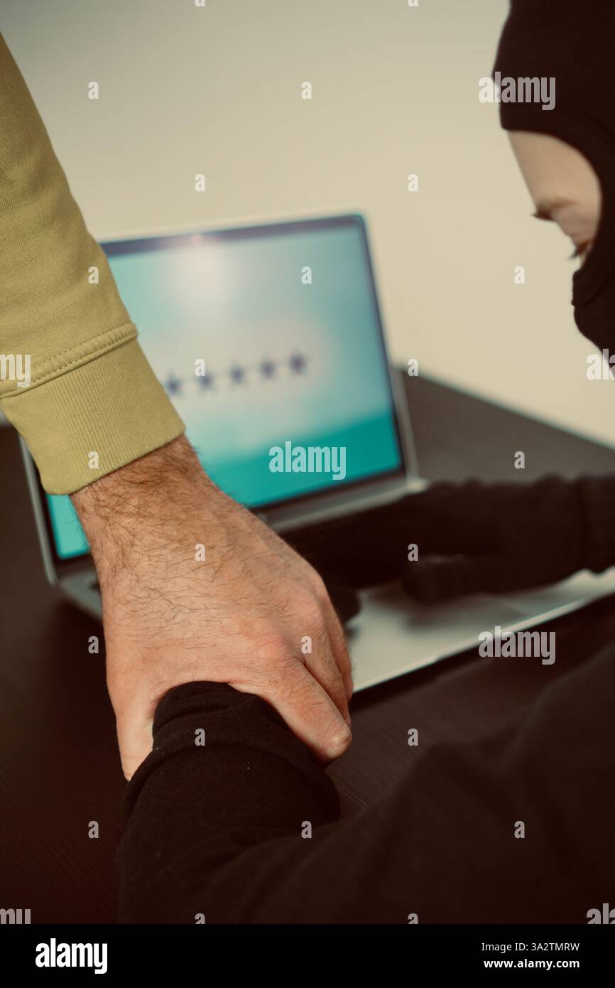 Un hacker in una balaclava nera ha colto la mano rossa che immette una password su un laptop, simboleggiando la criminalità informatica e le violazioni della sicurezza informatica. . Foto di alta qualità Foto Stock