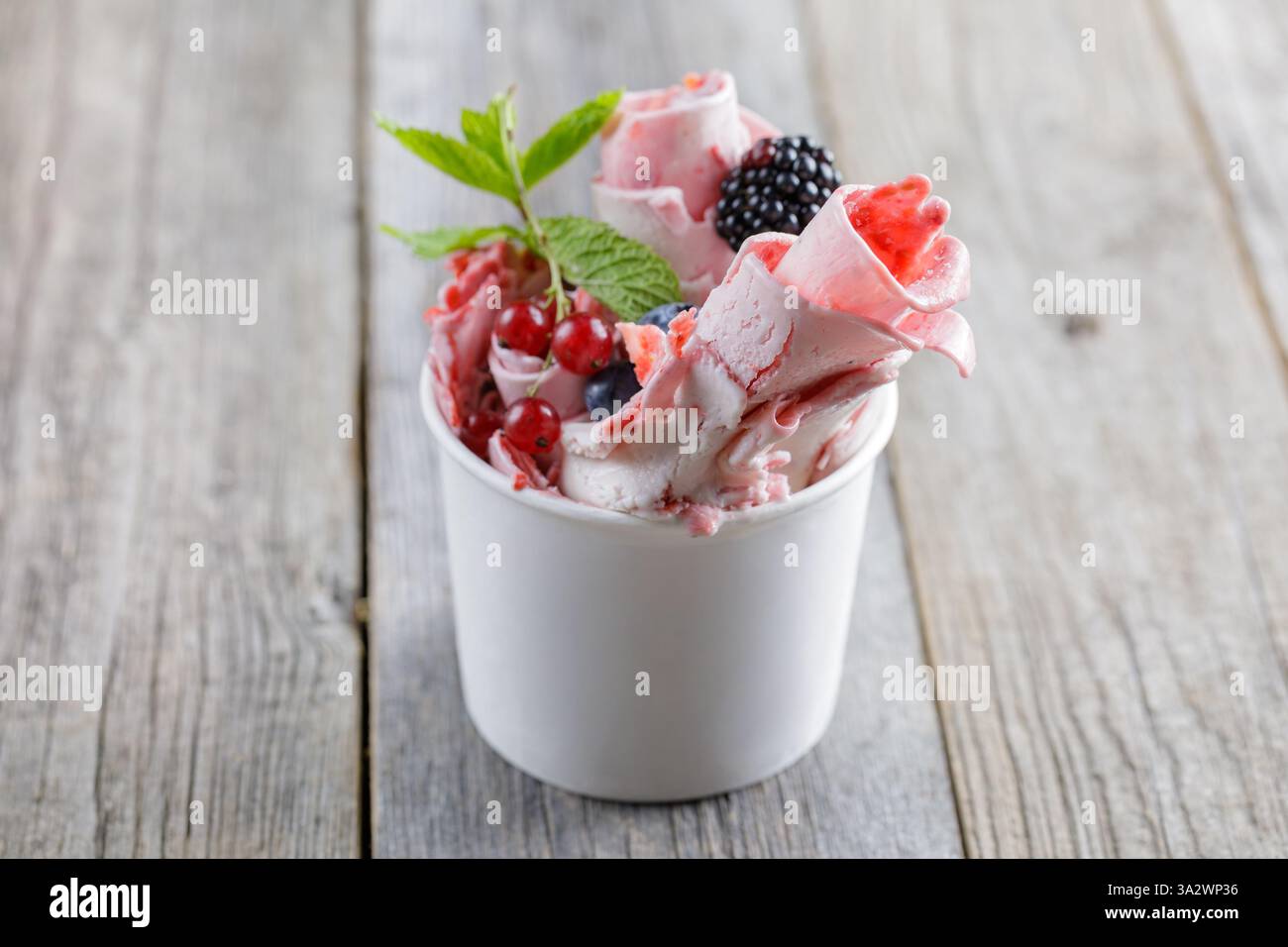 Delizioso gelato arrotolato in una tazza, guarnito con frutti di bosco freschi, un bastoncino di cioccolato e guarnizioni di frutta, servito su un rustico tavolo di legno Foto Stock