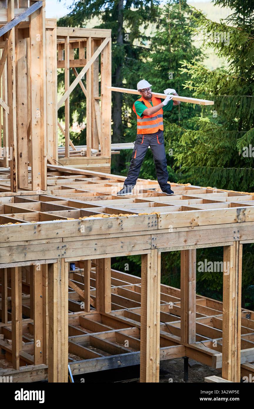 Edificio carpentiere casa con telaio in legno vicino alla foresta. Uomo barbuto con occhiali che reggono una grande tavola sulla spalla, vestito con abiti da lavoro e casco. Concetto di costruzione moderna e sostenibile. Foto Stock