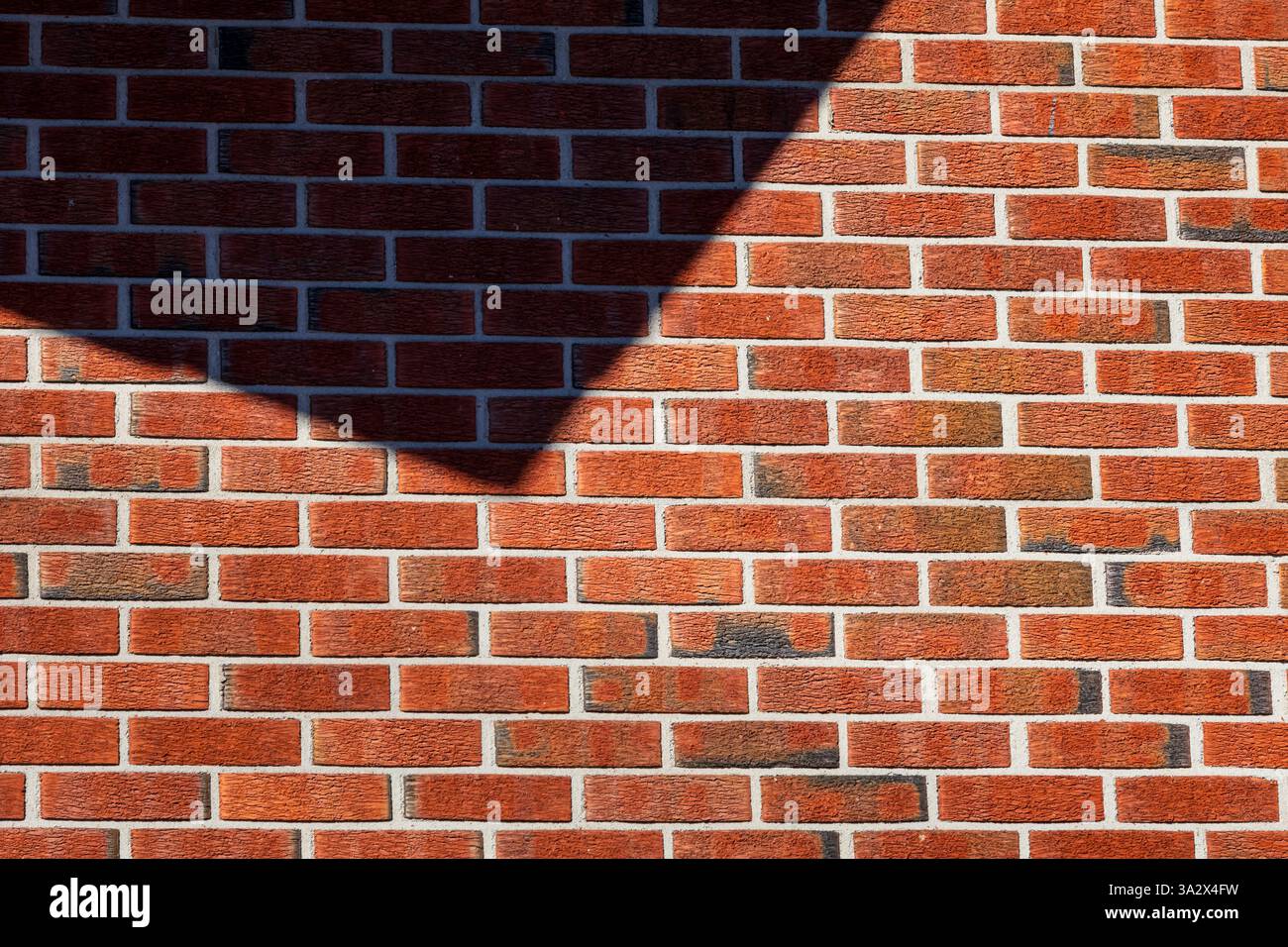 Un interessante gioco di ombre crea forme triangolari nitide su una parete di mattoni rossi, aggiungendo profondità e contrasto geometrico alla superficie ruvida Foto Stock