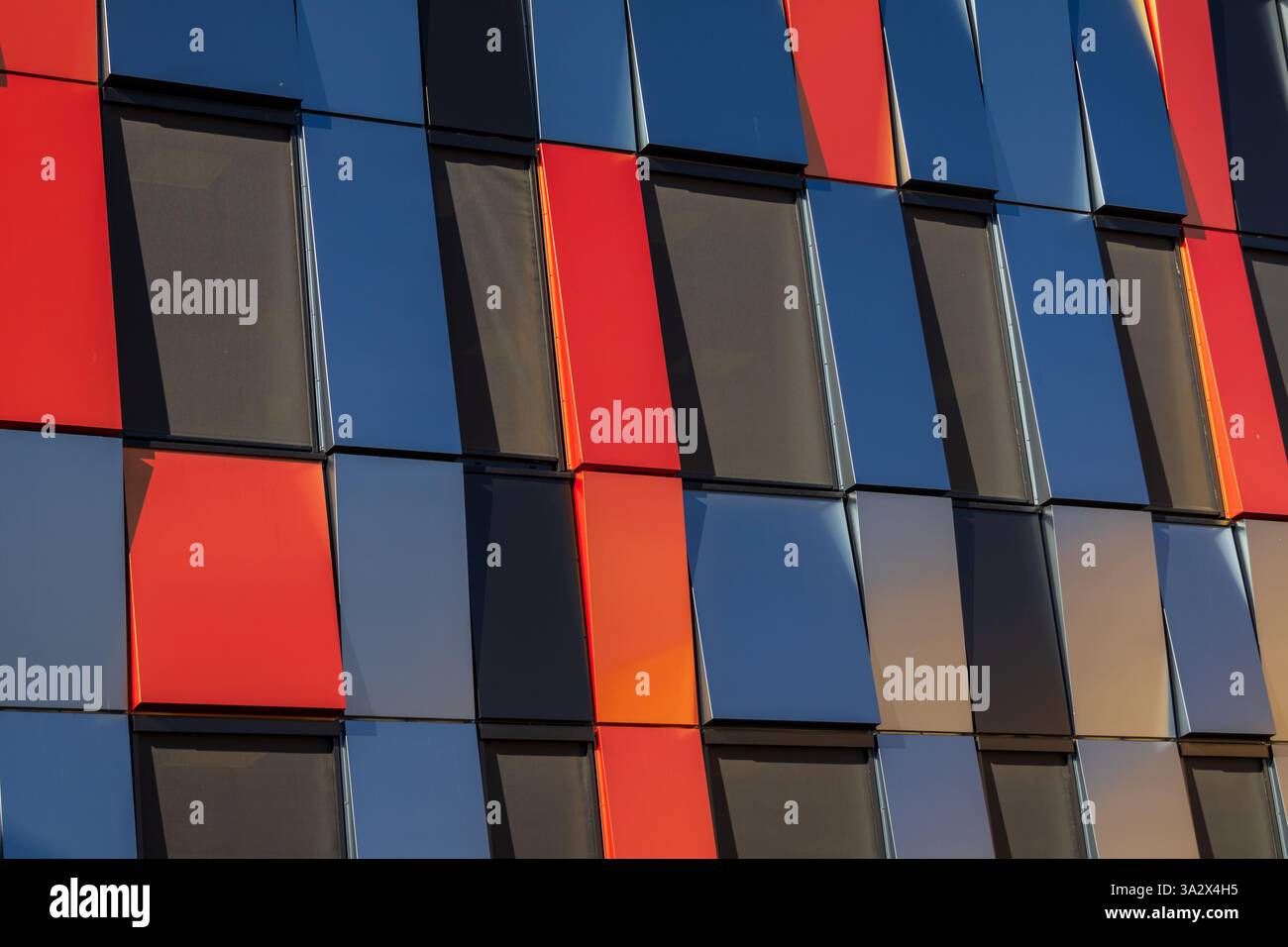 Una facciata moderna dell'edificio composta da rettangoli metallici rossi e blu scuro, sfalsati per rivelare schermi solari in tela nera, creando un contrasto dinamico Foto Stock