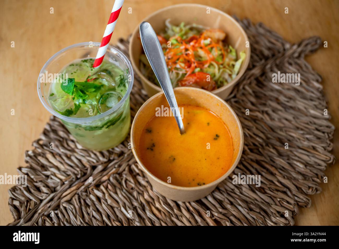 Zuppa piccante di cocco, insalata di verdure con gamberi in un recipiente di carta e cetrioli freschi con cannuccia su un cuscinetto di bambù sul tavolo. Cucina asiatica. Foto Stock