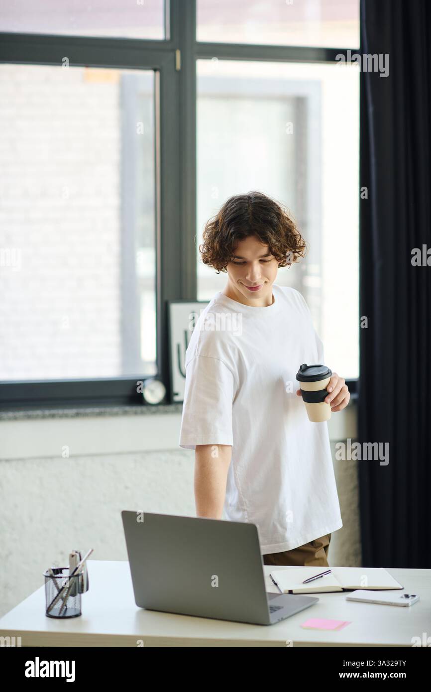 Un giovane attraente si trova in un ufficio luminoso, guardando il proprio laptop mentre si tiene una tazza di caffè. Foto Stock