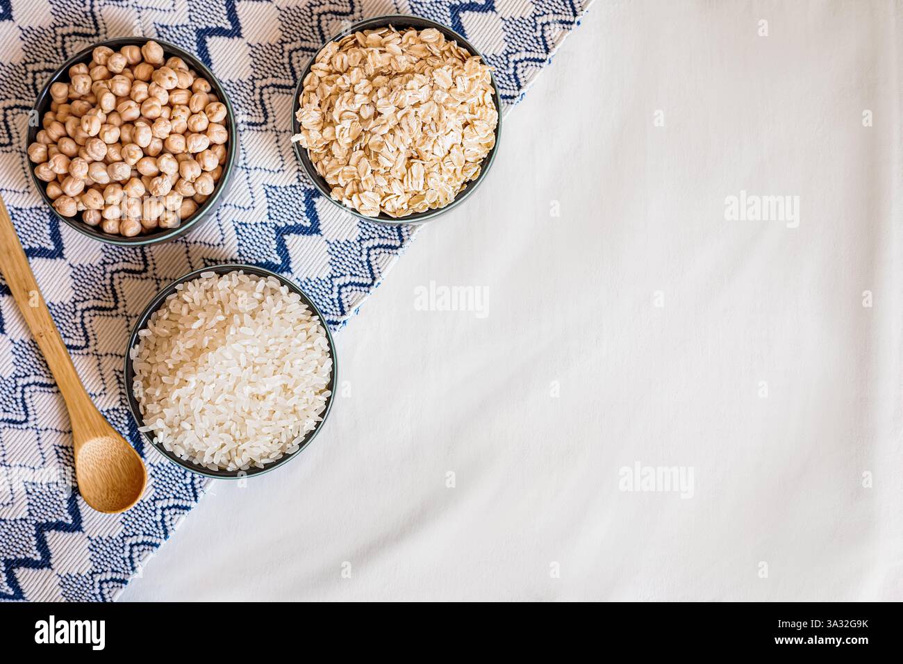 Vista dall'alto delle ciotole contenenti riso, ceci e avena, poste su un panno testurizzato con un cucchiaio di legno, evocano un'atmosfera culinaria accogliente e naturale Foto Stock
