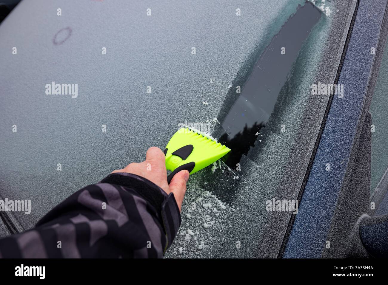 Mano maschile che usa un raschietto di plastica per ghiaccio sul finestrino di un'auto ghiacciata al mattino. Primo piano. Foto Stock