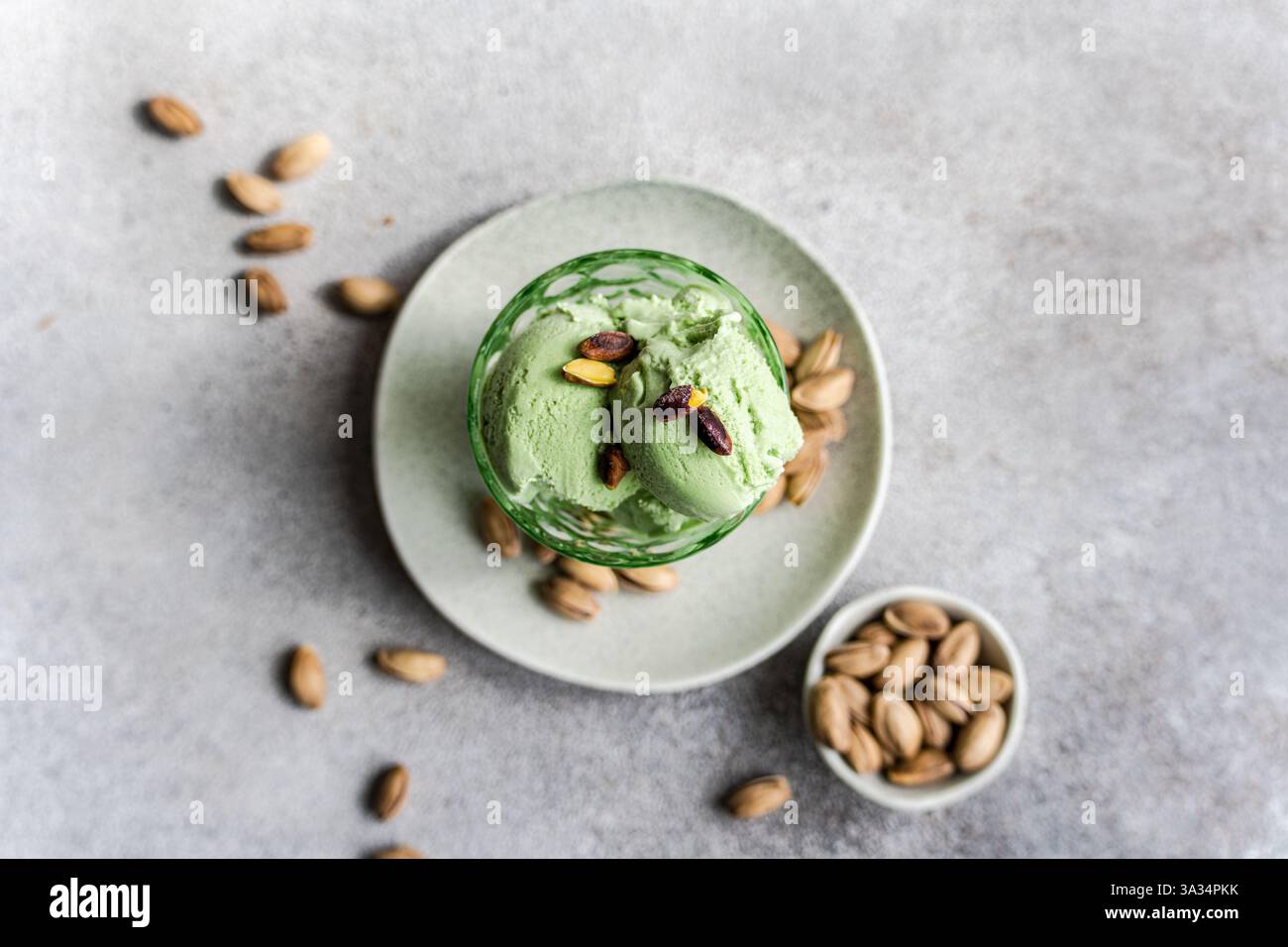 Due palline di gelato cremoso al pistacchio si trovano in una ciotola di vetro, guarnite con pistacchi interi. Il piatto viene posizionato su un piatto bianco con nocciole sparse Foto Stock
