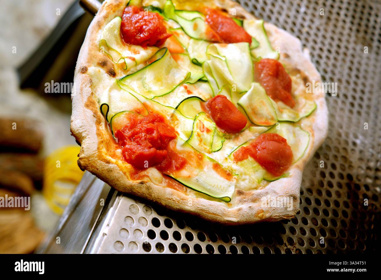 Una pizza vegetariana appena sfornata condita con fette di zucchine, pezzi di pomodoro, sollevati da una spatola da un vassoio per pizza perforato Foto Stock