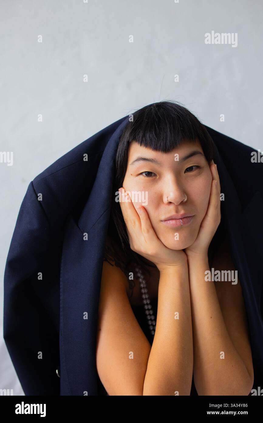 Un ritratto di una donna con un'acconciatura distintiva. La sua espressione facciale è premurosa, incorniciata da una giacca scura. Lo sfondo minimalista è complementare Foto Stock