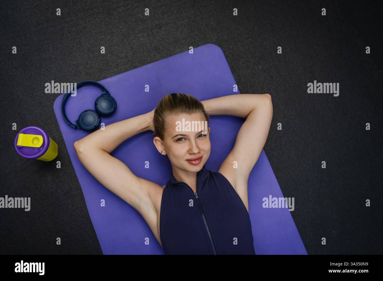 Una donna bionda in stile atletico si appoggia su un tappetino yoga viola con cuffie e una bottiglia d'acqua accanto a lei, a indicare il relax dopo un allenamento. Foto Stock
