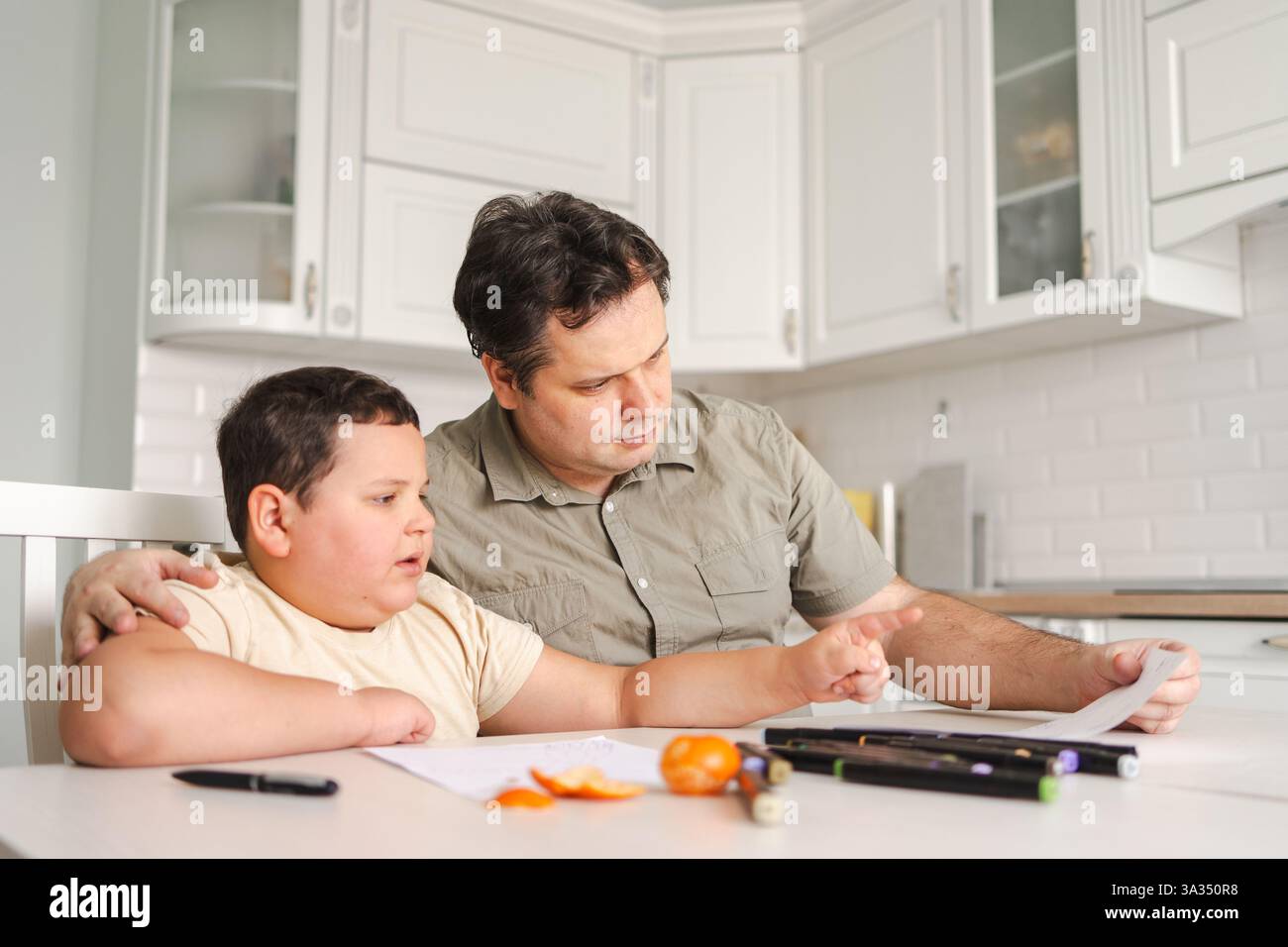 Un padre aiuta suo figlio con i compiti al tavolo della cucina. Sono circondati da forniture scolastiche, promuovendo un ambiente di apprendimento e legami familiari Foto Stock
