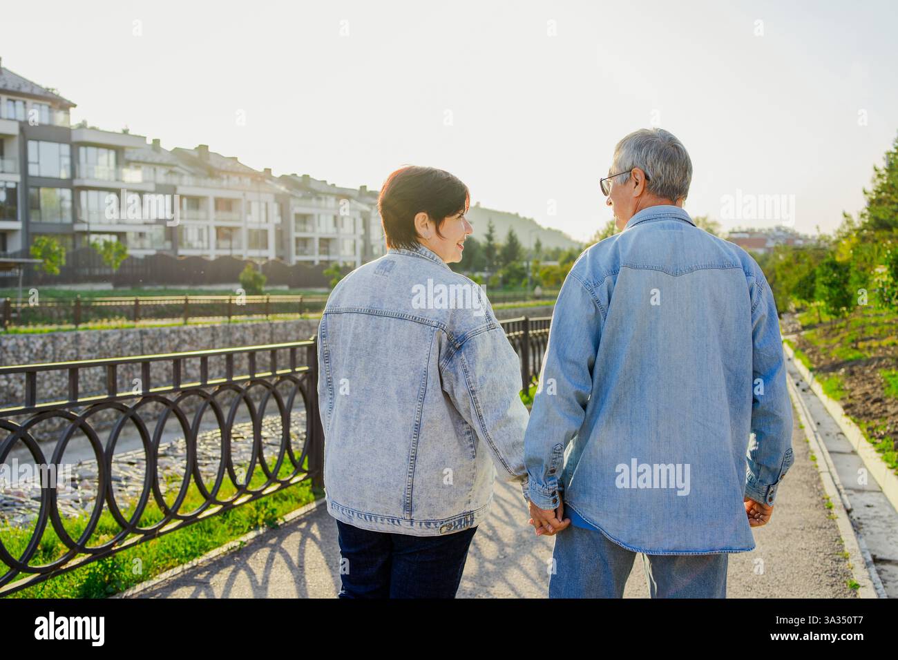 Una coppia di anziani cammina mano nella mano lungo un sentiero illuminato dal sole, indossando giacche in denim. Godono di un momento tranquillo vicino a un fiume, con appartamenti moderni Foto Stock