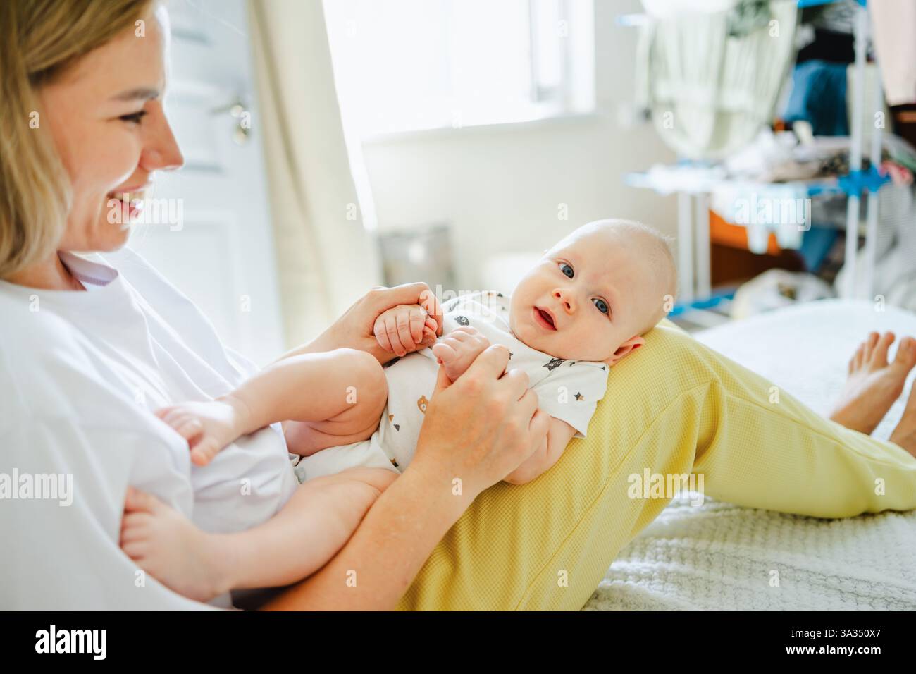 Una madre sorridente tiene il suo bambino in braccio a casa, godendosi preziosi momenti di legame. La camera luminosa aggiunge calore, enfatizzando l'amore materno e il conn Foto Stock