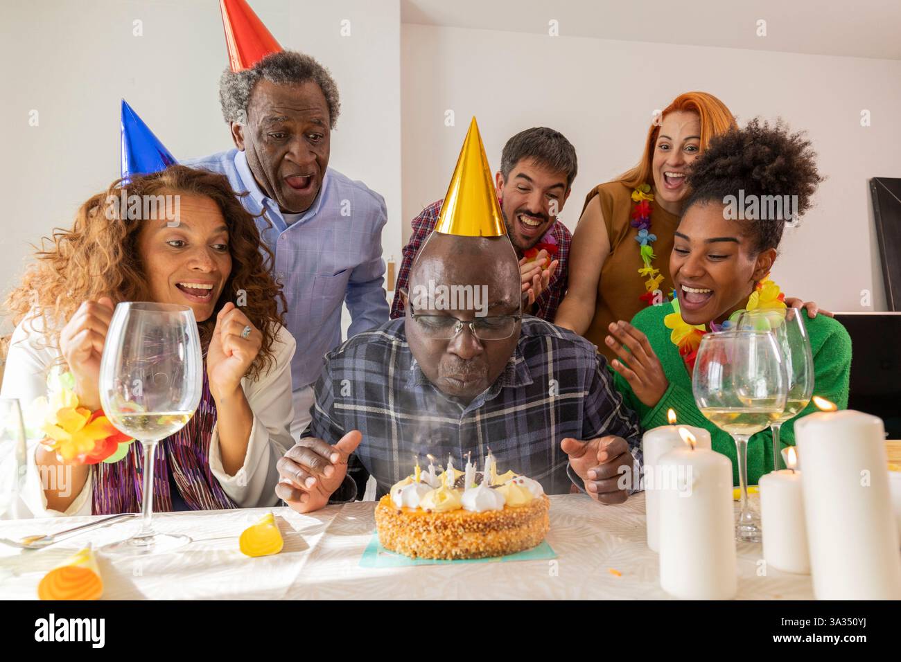 Un gruppo gioioso in occasione di una festa di compleanno. L'ospite d'onore indossa un cappello da festa, pronto a far esplodere le candele su una torta, circondato da amici Sha Foto Stock