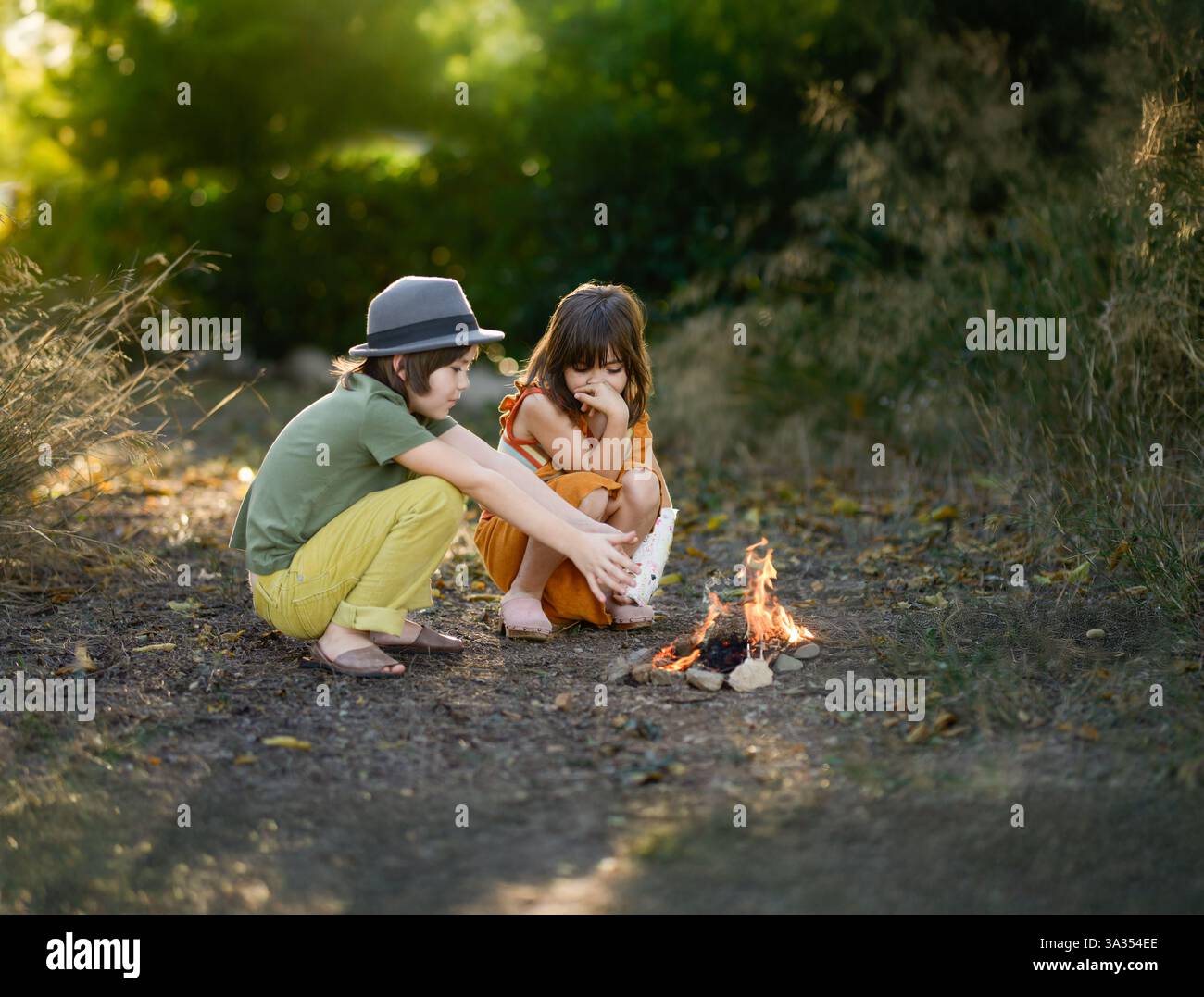 Due bambini siedono accanto a un piccolo falò in un'area boschiva. Si godono la vita all'aperto, circondati dalla natura. Il sole che tramonta crea un caldo e sgargiante Foto Stock