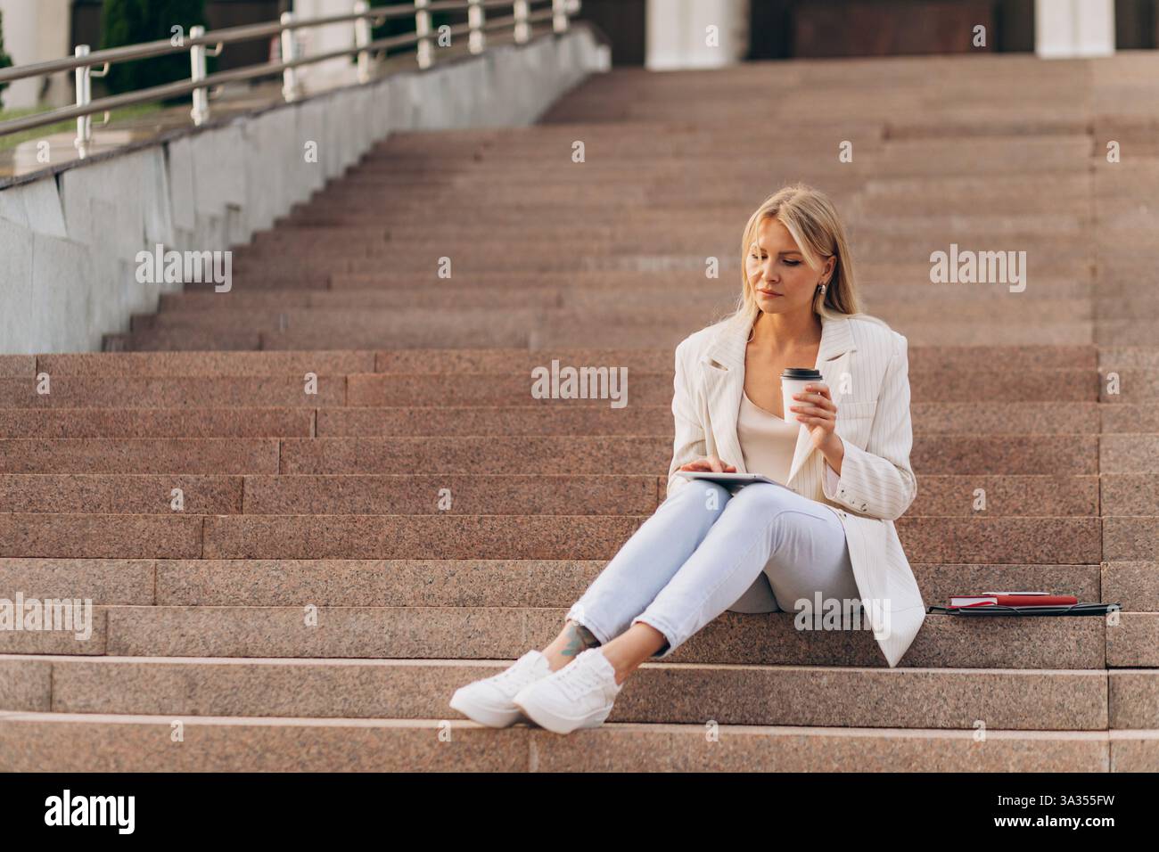 Una donna d'affari con un abbigliamento casual si siede sui gradini all'aperto, tenendo una tazza di caffè e usando un tablet. Un momento di serenità che mette in risalto l'equilibrio tra Foto Stock