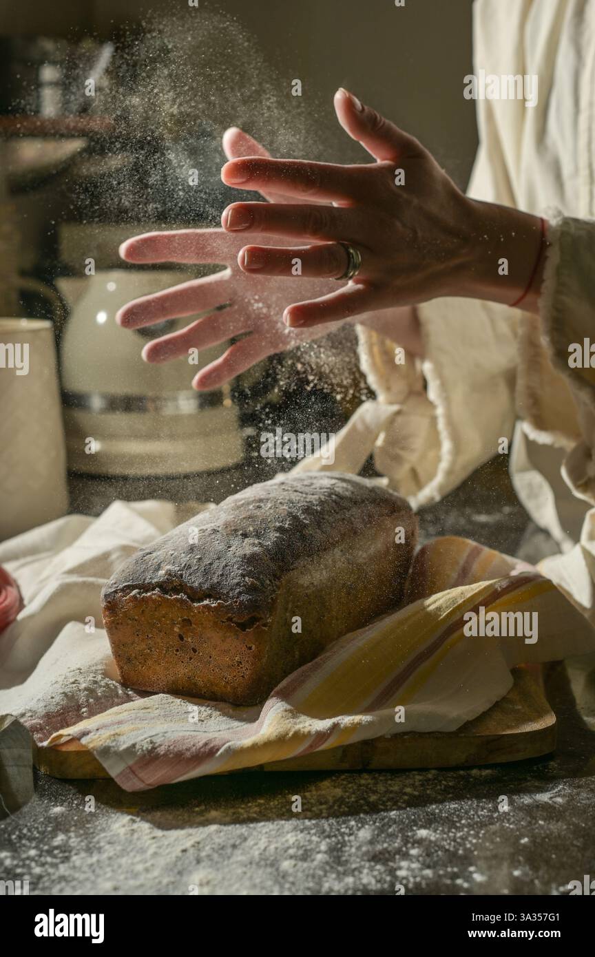 Una scena calda e rustica di cottura del pane. Le mani applaudono per spolverare la farina mentre una pagnotta di pane si raffredda su un tagliere di legno, catturando un'immagine minimalista e. Foto Stock