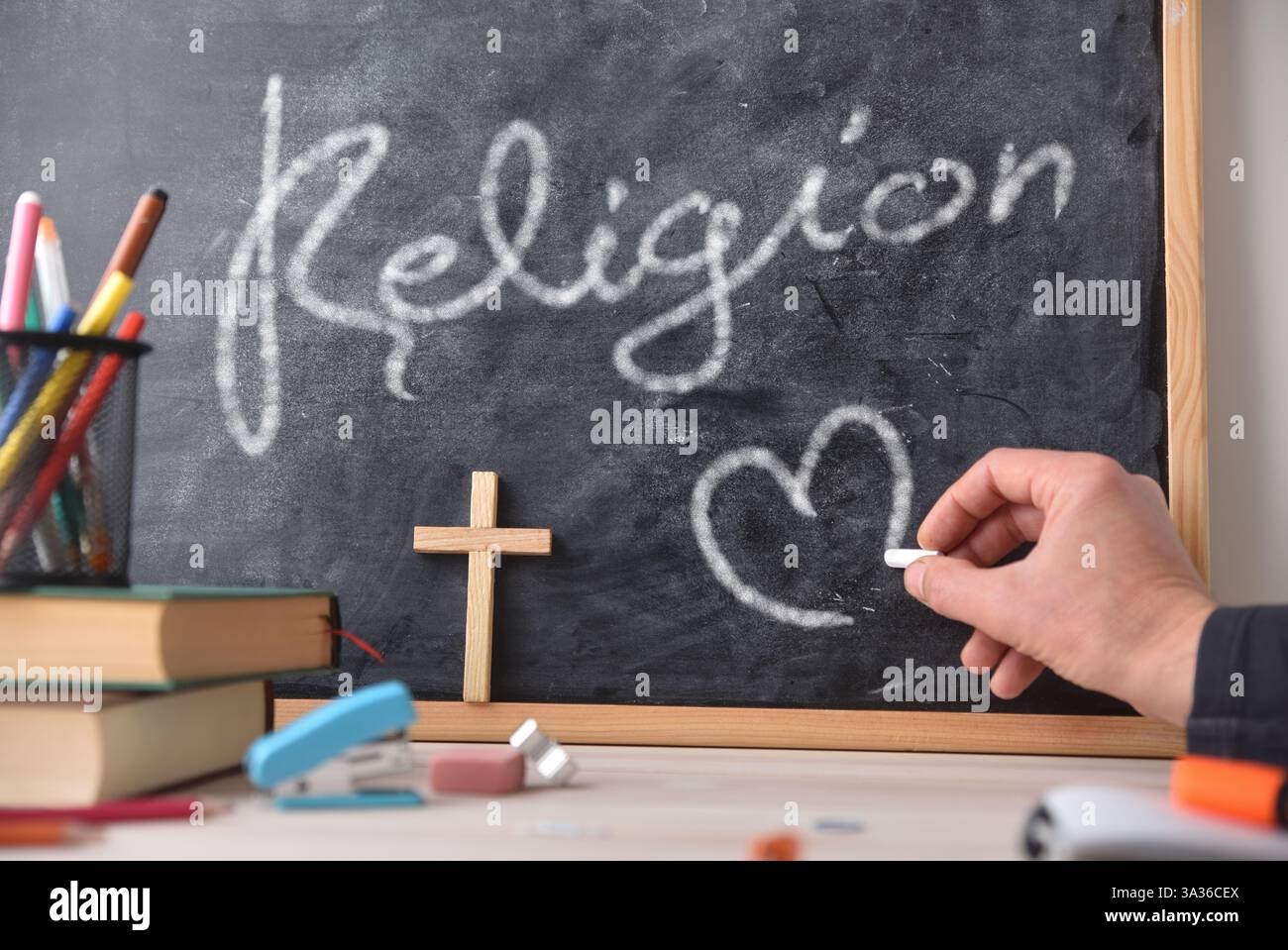 Disegnare a mano un cuore con il gesso su una lavagna con la parola religione scritta sopra. Una scrivania di legno con libri e materiale scolastico. Vista frontale. Foto Stock