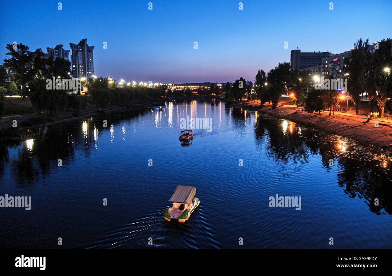 Le acque calme riflettono le luci della città mentre le barche navigano sul fiume sereno, creando un'atmosfera tranquilla al crepuscolo dagli sviluppi urbani. Foto Stock