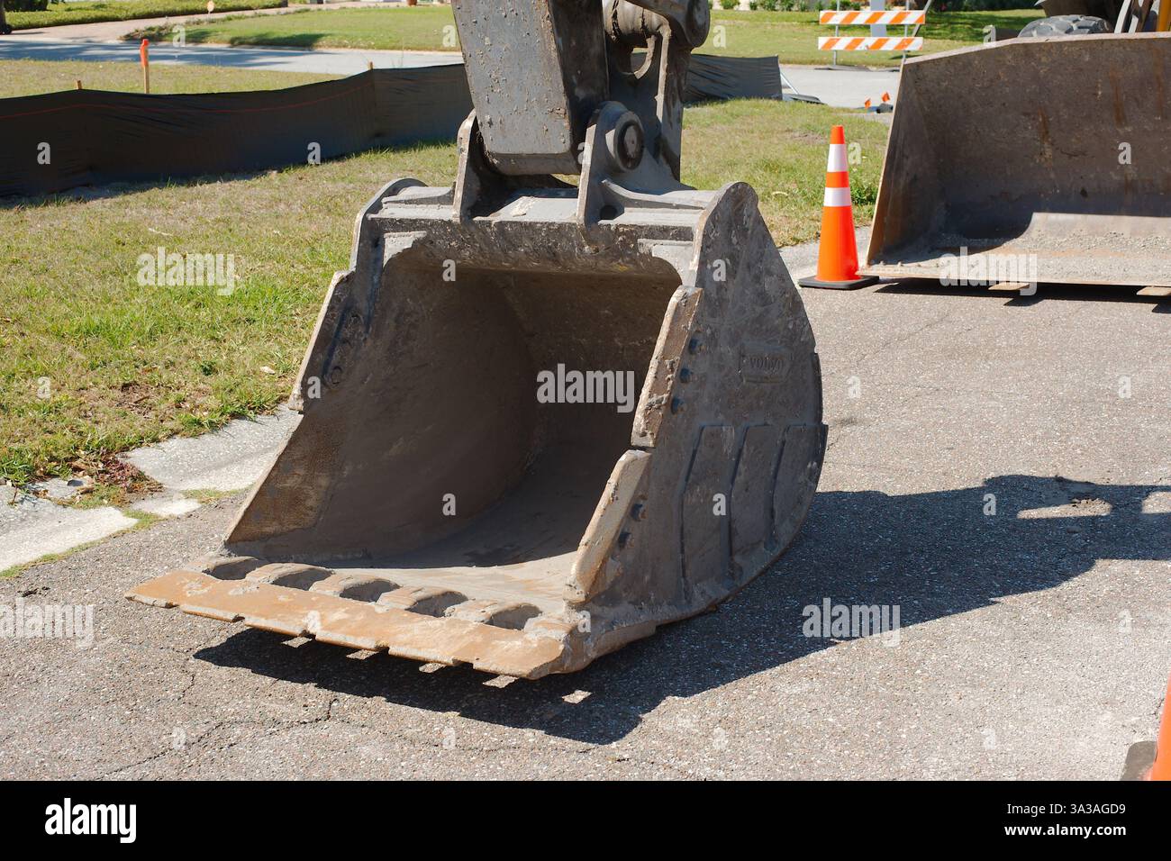 Primo piano di una benna dell'escavatore in acciaio, con denti visibili lungo il bordo per lo scavo. È posizionato su una superficie pavimentata con una struttura Foto Stock