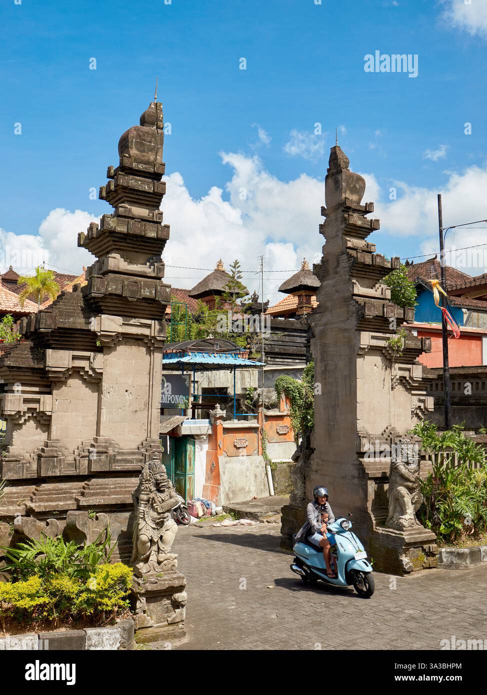 Una donna che tiene in braccio il suo bambino pedala in moto blu attraverso candi bentar, una classica porta balinese divisa. Distretto di Mengwi, Bali, Indonesia. Foto Stock