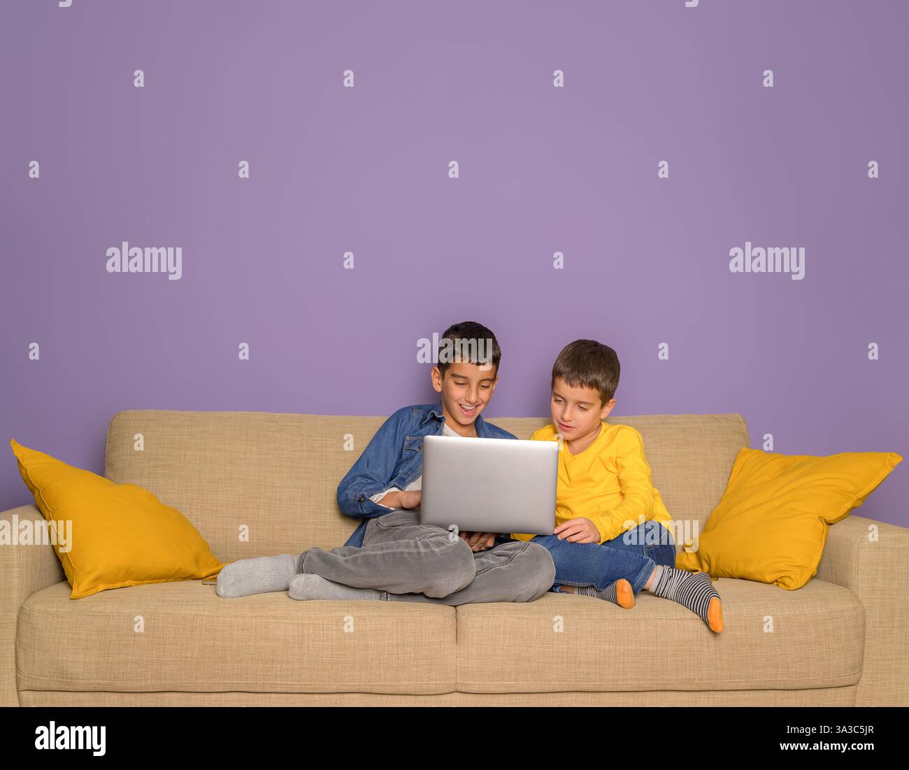 Giovani fratelli felici che guardano film su un notebook mentre si rilassano sul divano su sfondo viola Foto Stock