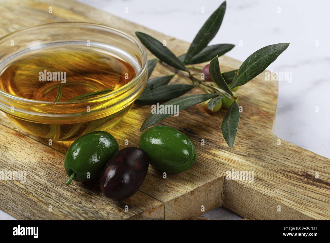 Una ciotola di vetro piena di ricco olio d'oliva si trova su un tagliere di legno accanto a olive verdi e nere fresche, completate da foglie d'oliva, creando Foto Stock