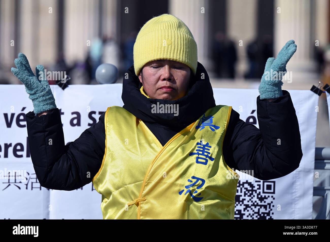 Piazza degli Eroi, Budapest, Ungheria - 2 marzo 2025: Manifestante del Falun Gong con giacca gialla e cappello che meditano in posizione eretta del Falun Foto Stock