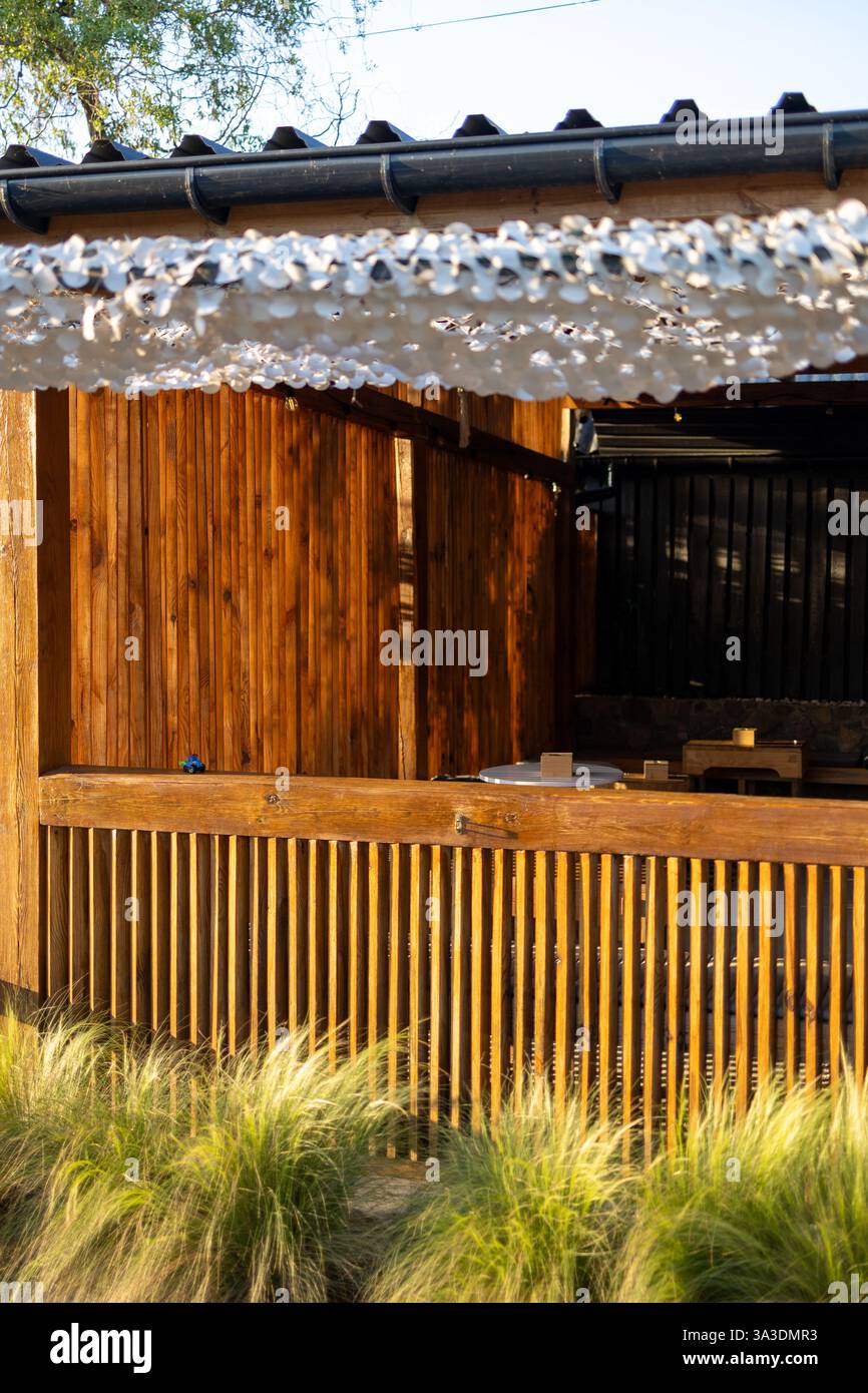Un bar rustico in legno con pannelli a doghe è inondato dalla calda luce del sole. Una rete decorativa bianca è appesa sopra, creando ombre morbide. Erba alta nella parte anteriore Foto Stock
