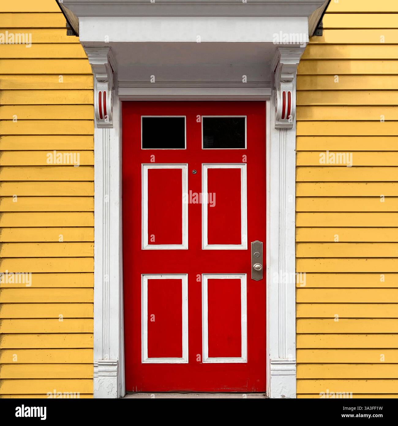 Porta rossa vibrante con finiture bianche adagiata su una facciata laterale in legno giallo, in un ambiente urbano. - Immagine stock catturata con smartphone
