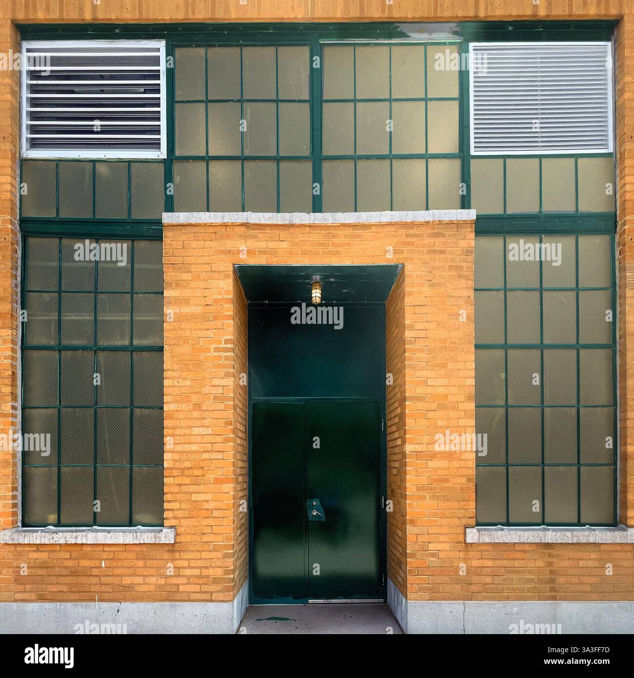 Ingresso all'edificio industriale caratterizzato da una porta in metallo verde incassata all'interno di una facciata in mattoni gialli, incorniciata da grandi finestre a griglia con pannelli di ventilazione. Foto Stock