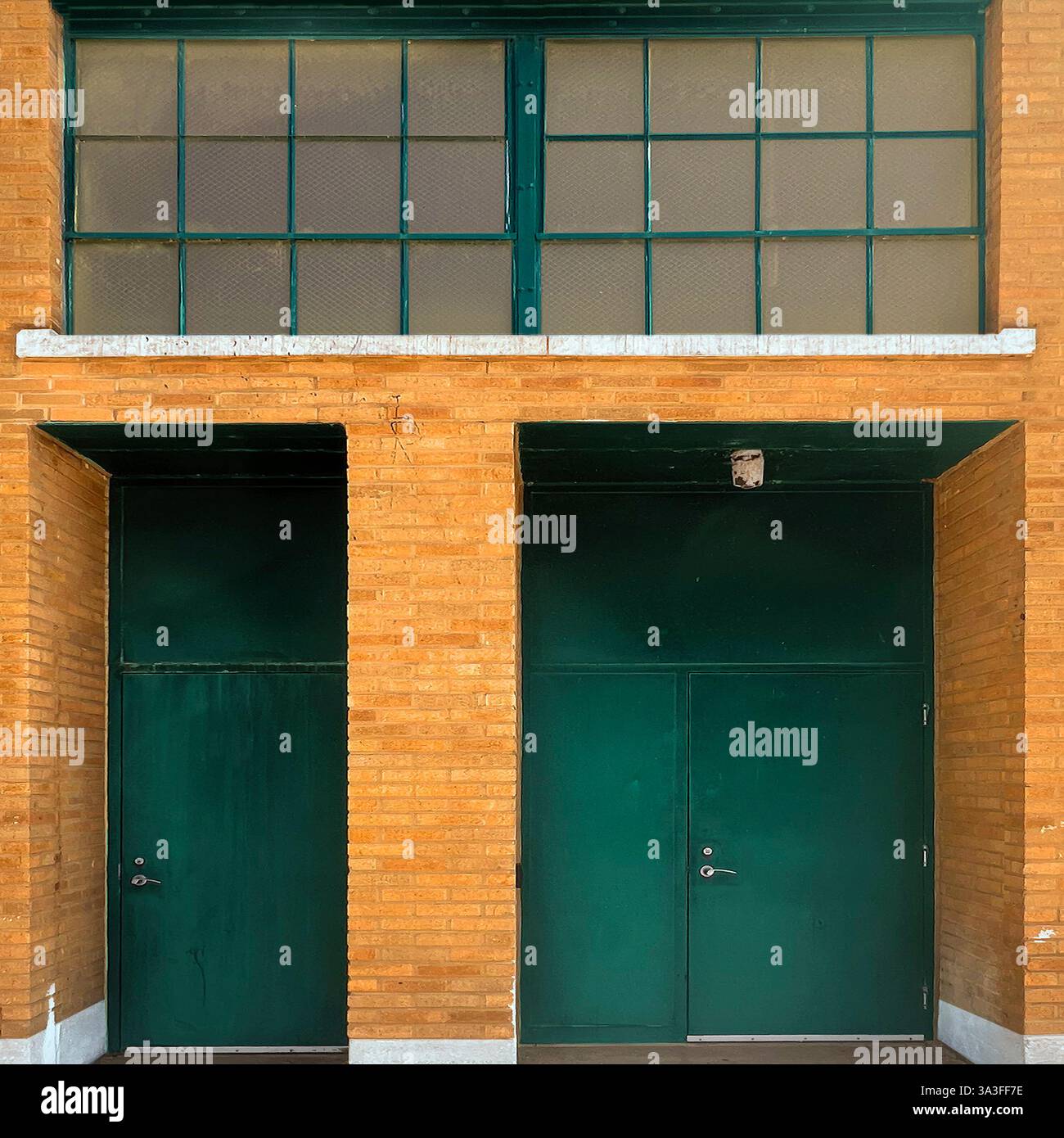 Ingresso all'edificio industriale con due porte di metallo verde scuro poste all'interno di una facciata in mattoni gialli, finestre a griglia incorniciate e una luce di sicurezza superiore. Foto Stock